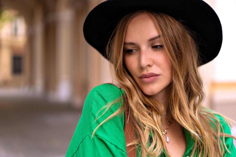 Close up portrait of elegant stylish woman with blonde curly hair and natural make up wearing black fedora posing on the street
