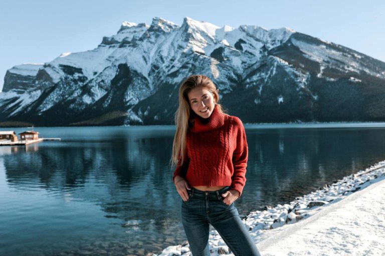 Pretty lady with white smile standing on the beach near the lake. mountains covered with snow. wearing red knitted sweater and blue jeans. blonde long hairstyle no makeup
